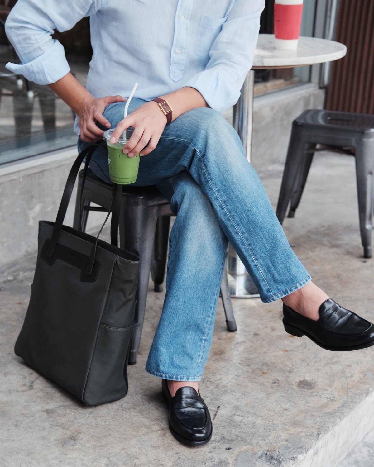 Person wearing a light blue shirt, blue jeans, and black loafers sitting outdoors.