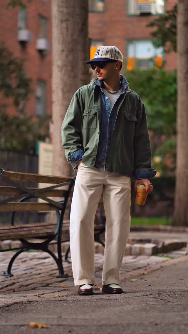 Man wearing olive jacket, denim shirt, and cream trousers standing in a park.