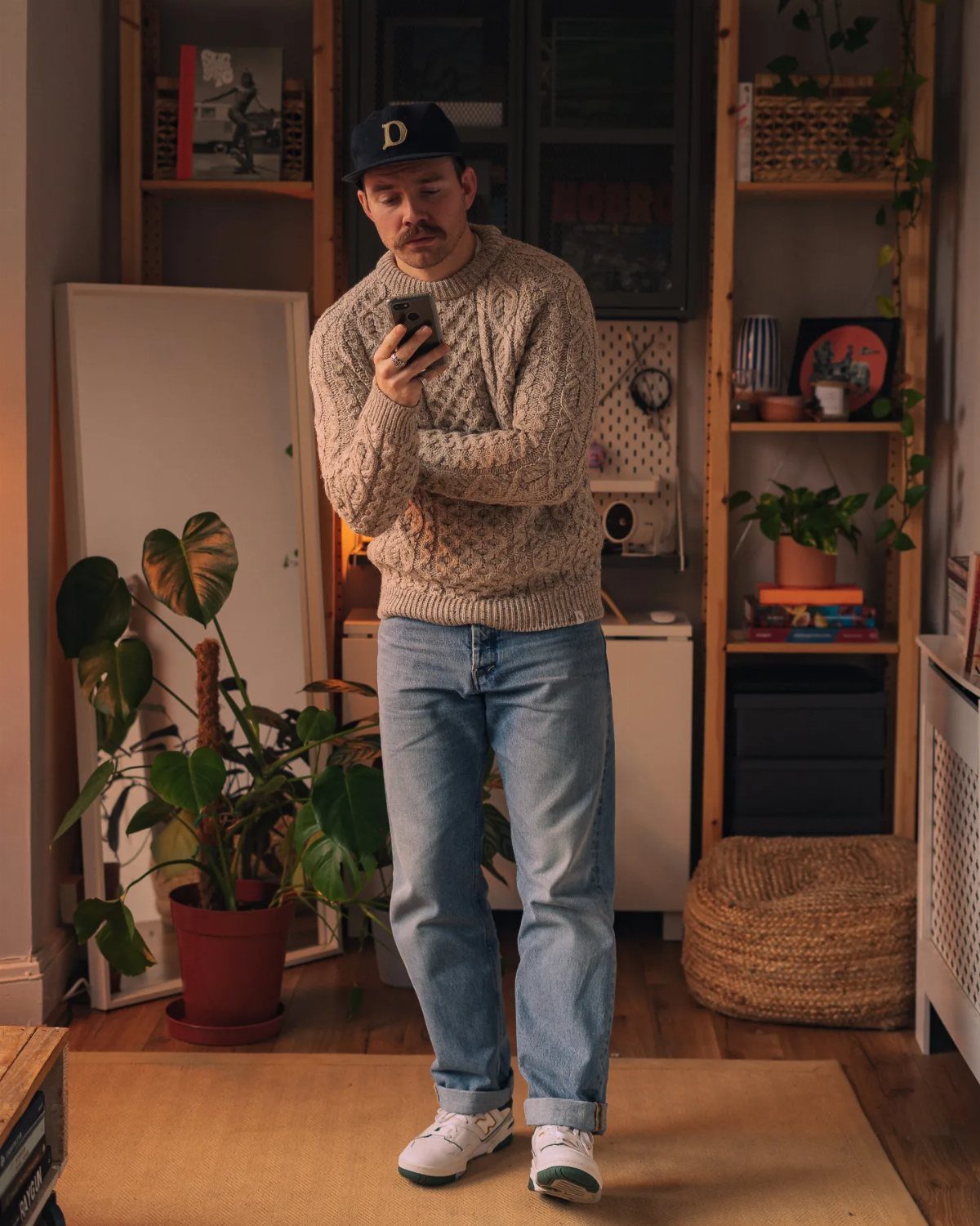Man in beige knit sweater and wide-leg jeans looking at his phone indoors.