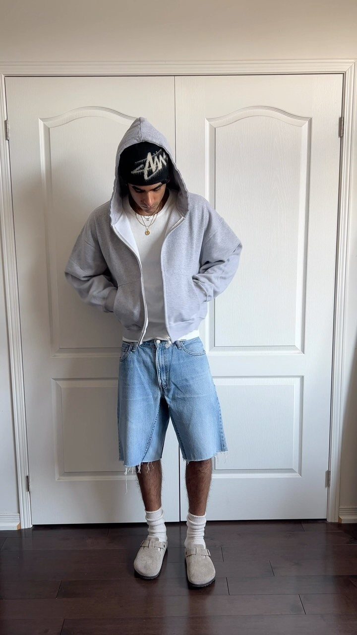 Man wearing a grey hoodie, white t-shirt, denim shorts, and beige shoes.