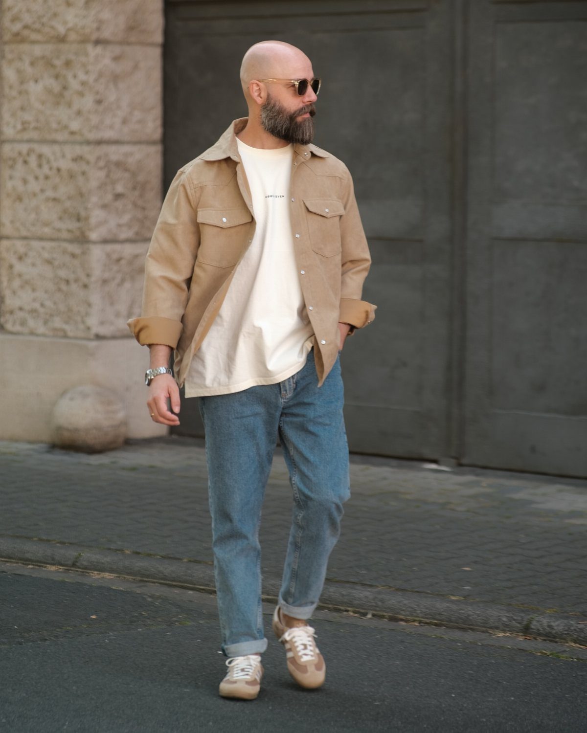 Man wearing a tan overshirt, cream t-shirt, blue jeans, and tan sneakers outdoors.