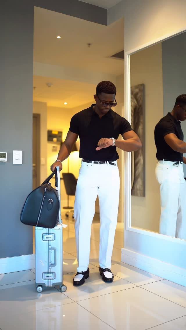 Man in black polo and white trousers standing with a silver suitcase.