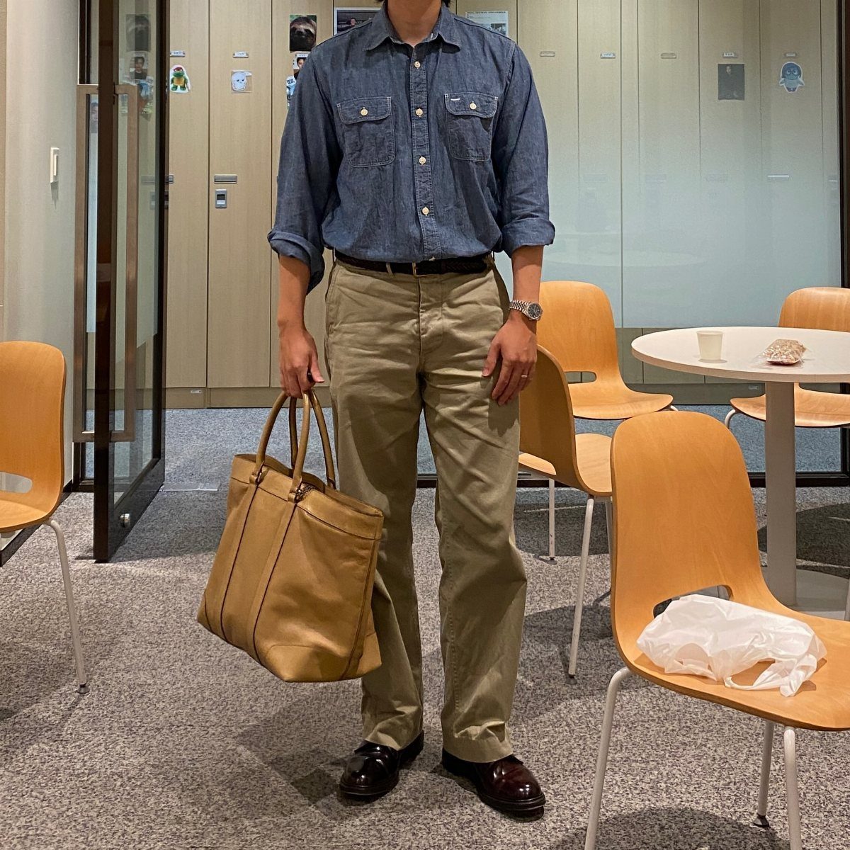 Person wearing a blue button-down shirt and khaki trousers holding a tan tote bag.
