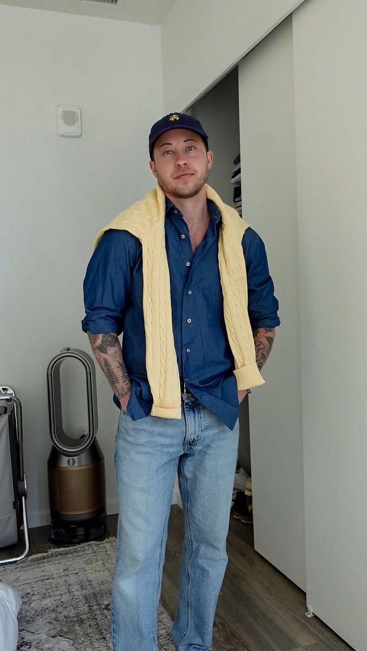 Man wearing a blue button-down shirt, light jeans, and yellow sweater over shoulders.