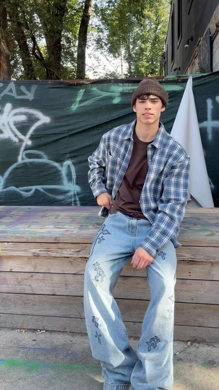 Man in a blue plaid flannel shirt, brown beanie, and wide-leg jeans sitting outdoors.