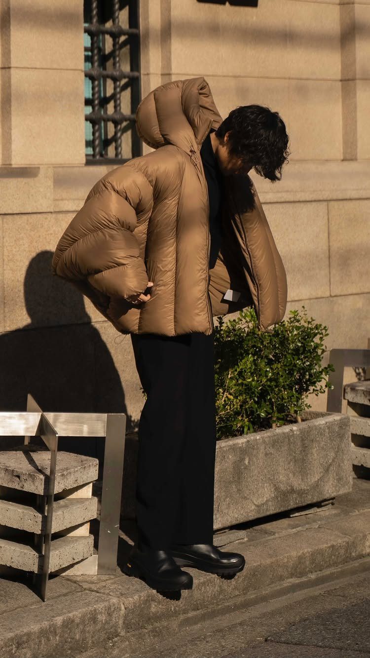Person wearing an oversized tan puffer jacket and black trousers outdoors by a building.