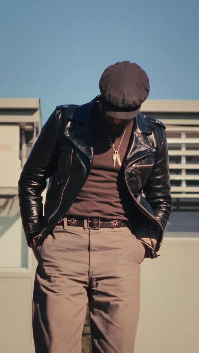 Person wearing a black leather jacket, tan trousers, and a black newsboy cap.