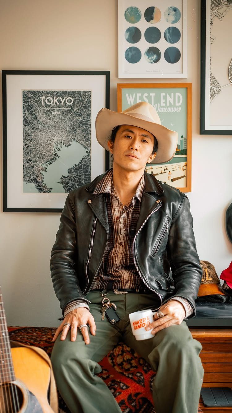Man in black leather jacket, cowboy hat, and olive pants sitting indoors.