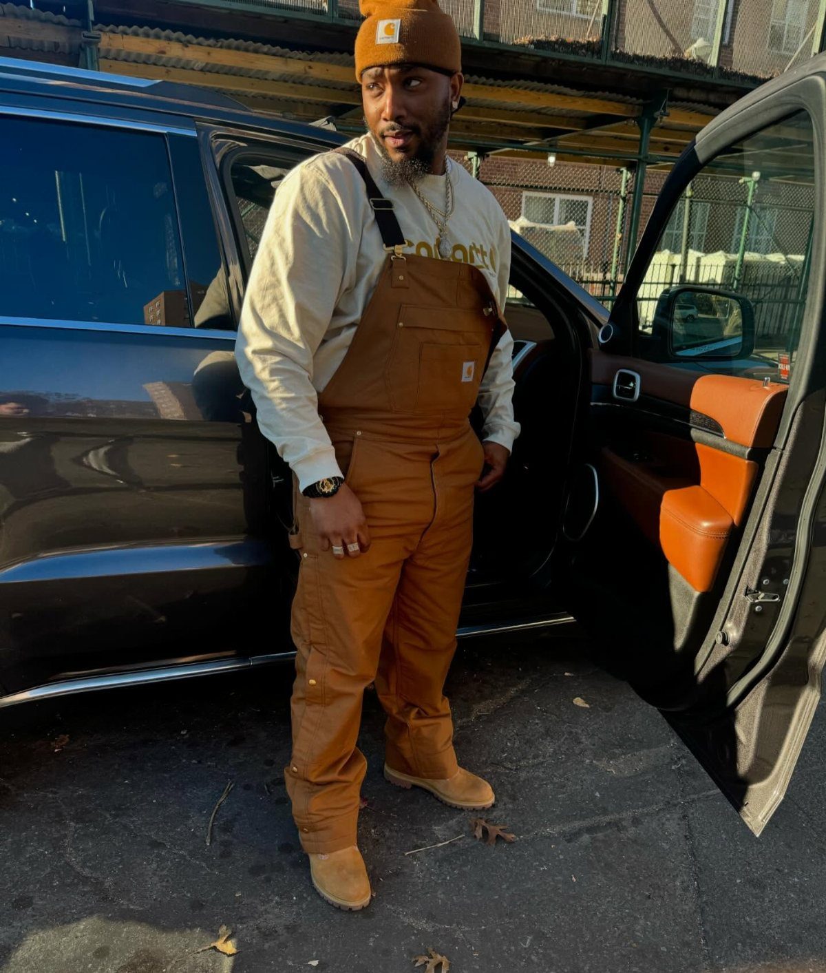 Man in brown baggy overalls and a brown beanie standing by a car.