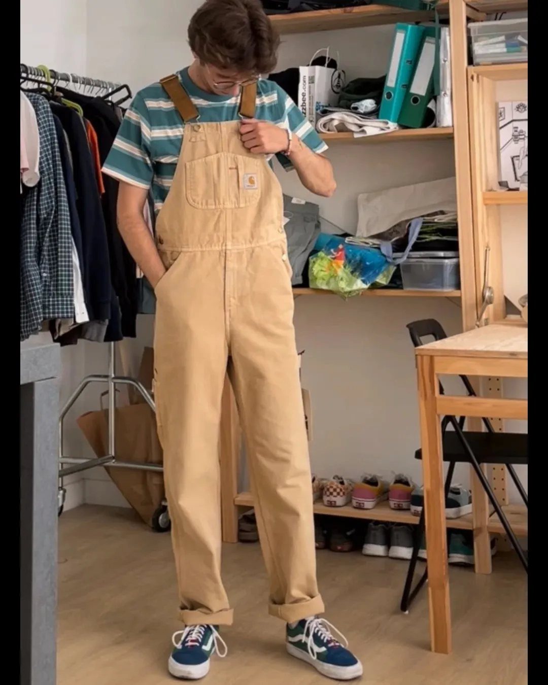 Person wearing tan baggy overalls and blue sneakers in a room with shelves.