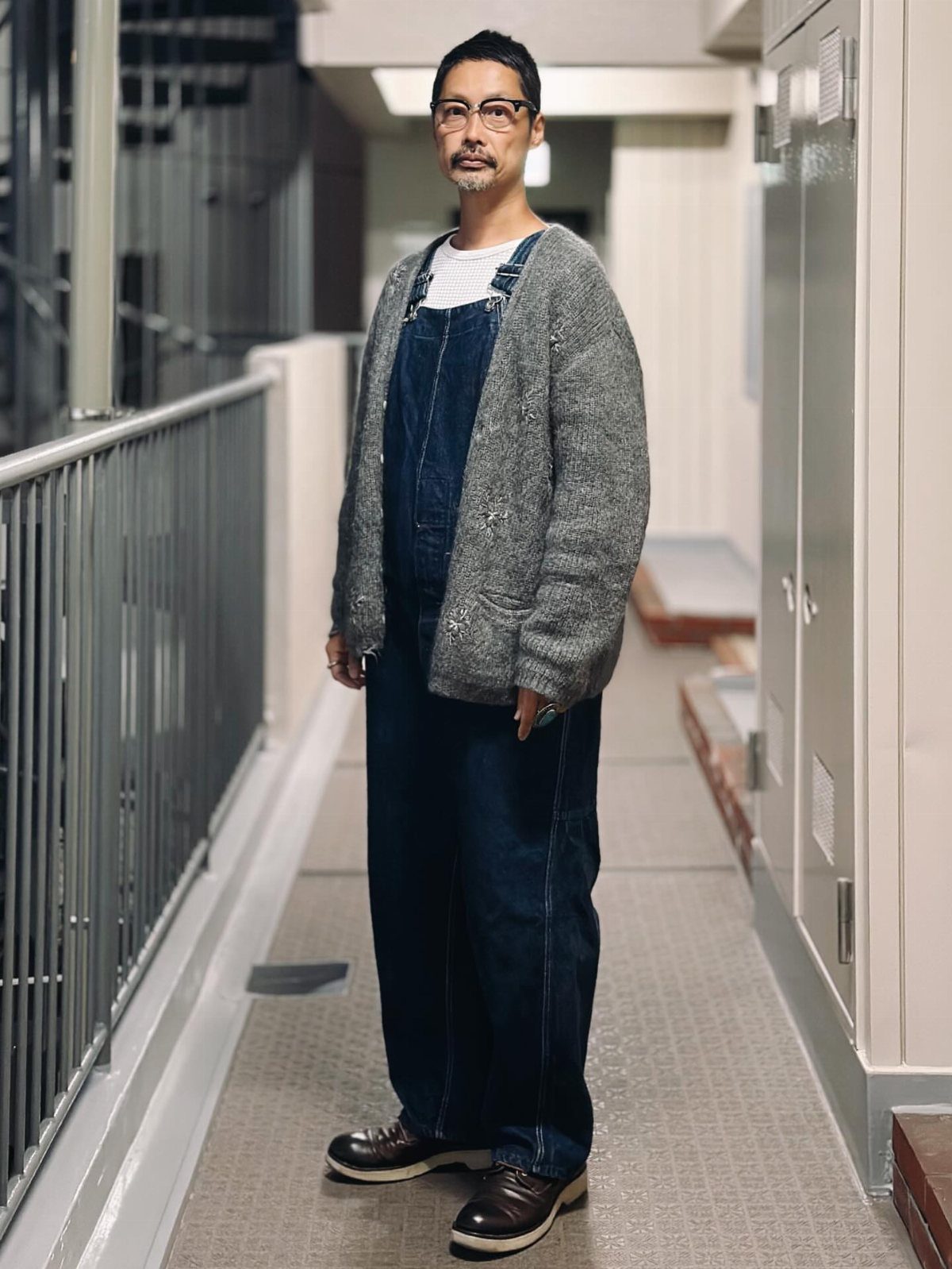 Man wearing a grey cardigan over blue denim baggy overalls in a hallway