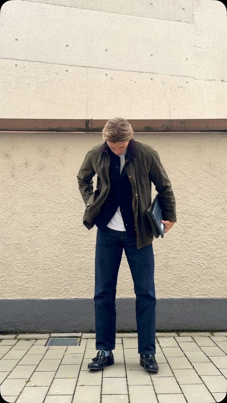 Man wearing an olive green Barbour jacket, navy cardigan, and dark blue jeans.