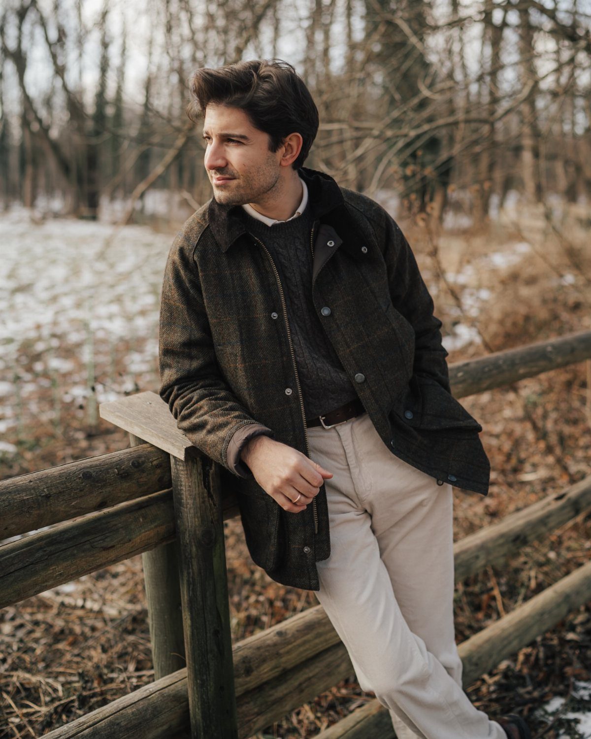 Man wearing a brown Barbour jacket and grey knitwear leaning on a fence