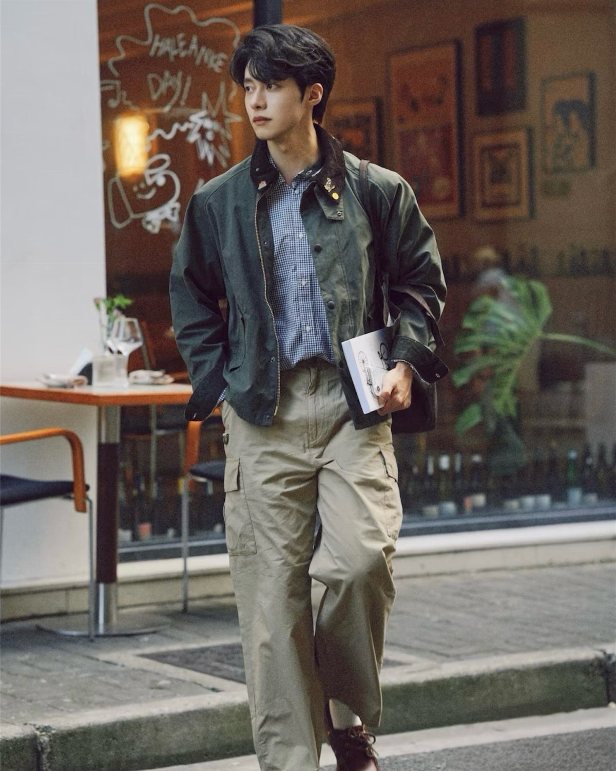 Man wearing a green Barbour jacket, checkered shirt, and beige cargo trousers walking outdoors.