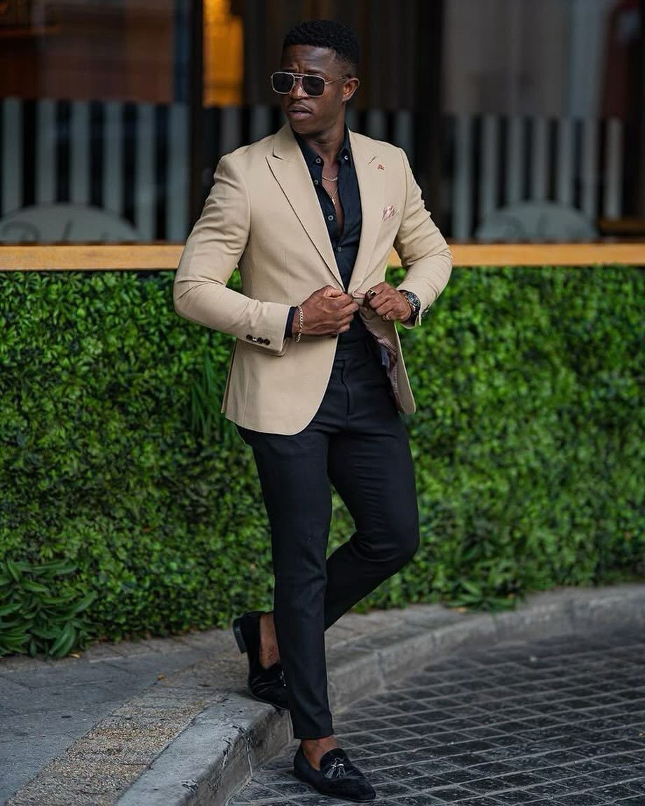 Man in a beige blazer and tailored black trousers standing by a green hedge.