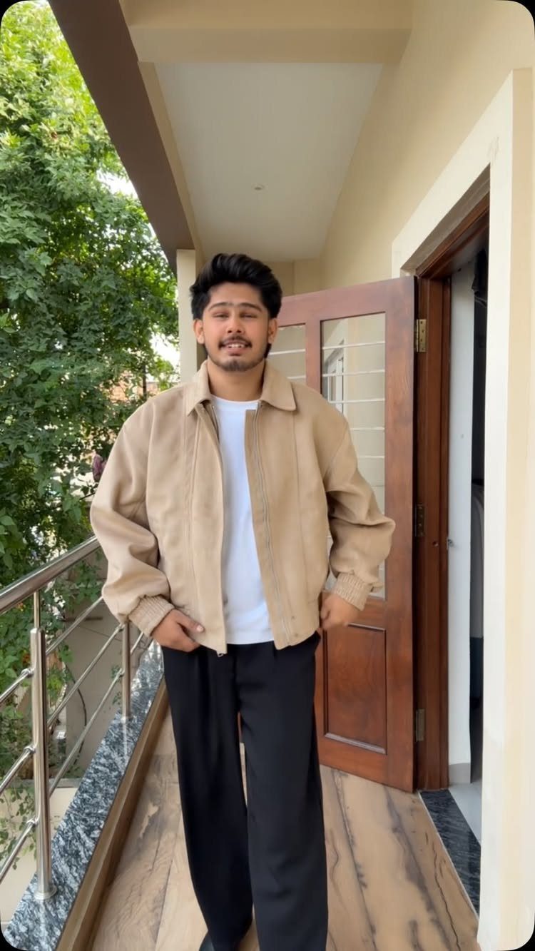 Man wearing a beige jacket, white t-shirt, and black trousers on a balcony.