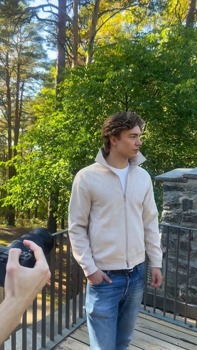 Man wearing a beige jacket and blue jeans standing on a wooden deck.