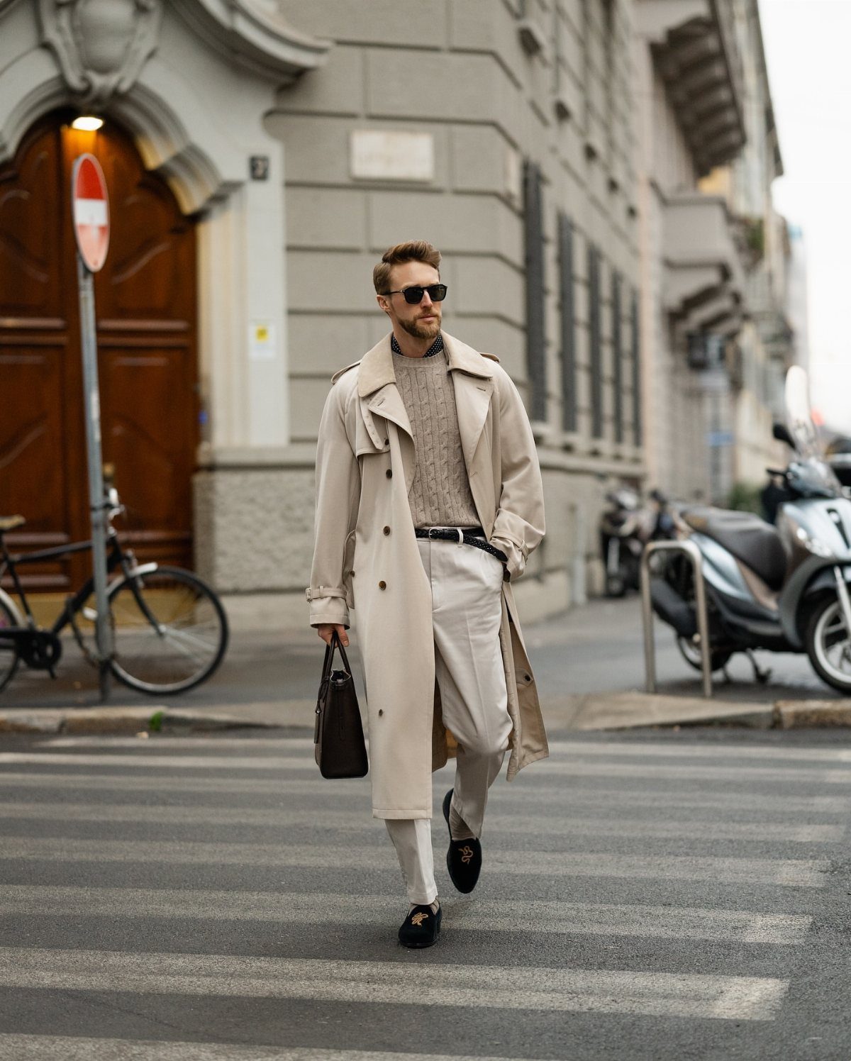 Man in a beige trench coat and white trousers walking across a city street