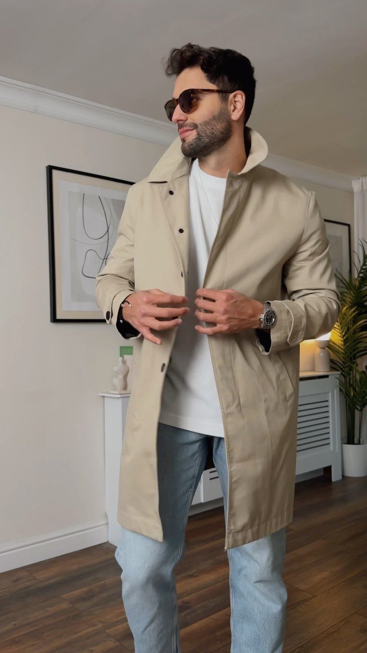 Man wearing a beige trench coat, white t-shirt, and light blue jeans indoors.