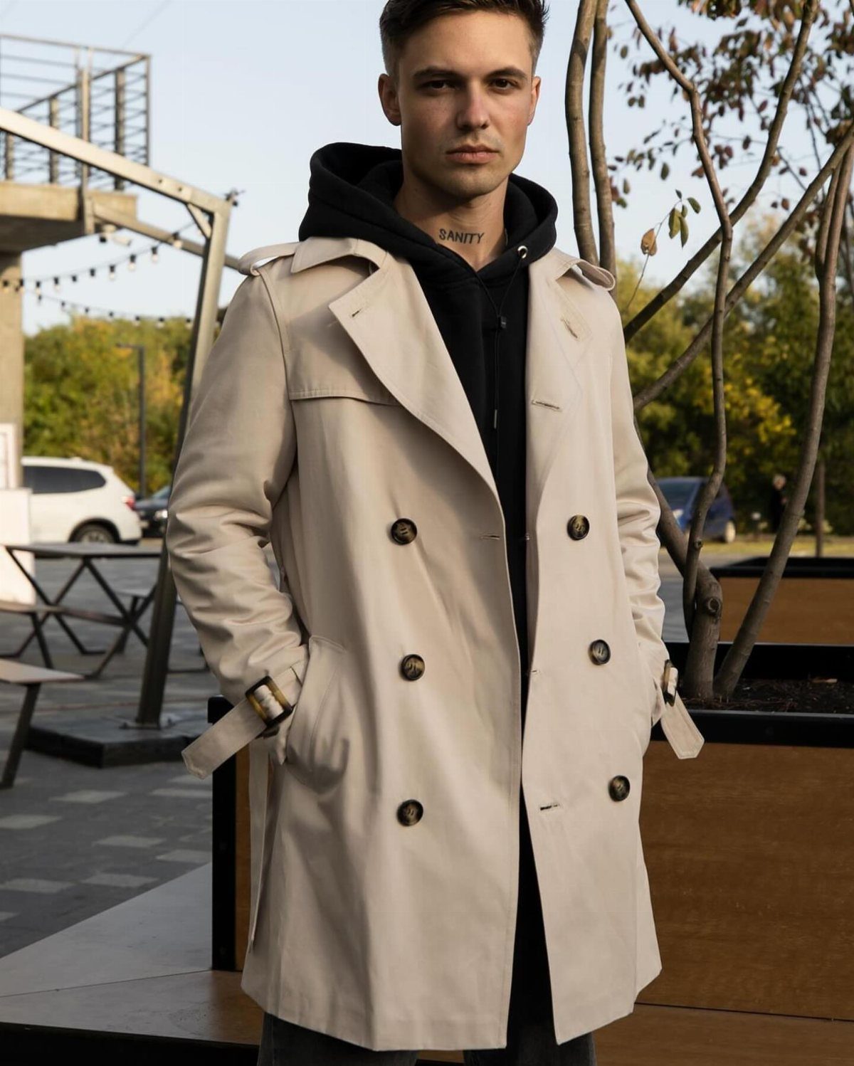 Man wearing a beige double-breasted trench coat over a black hoodie outdoors.