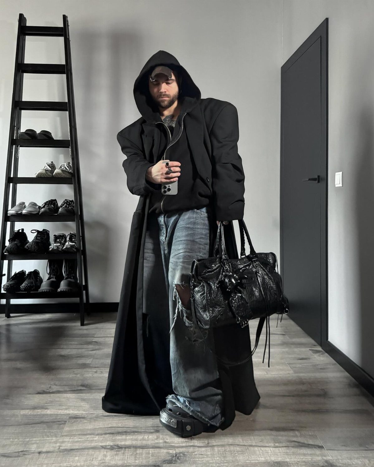 Man in a long black hooded coat and ripped blue jeans holding a handbag.