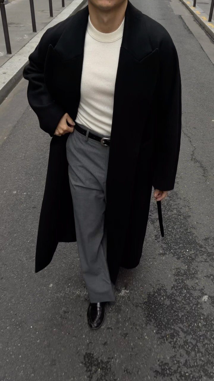 Person wearing an oversized long black coat with grey trousers and black shoes.