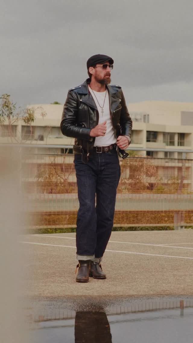 Man in black leather jacket and blue jeans wearing a black flat cap.