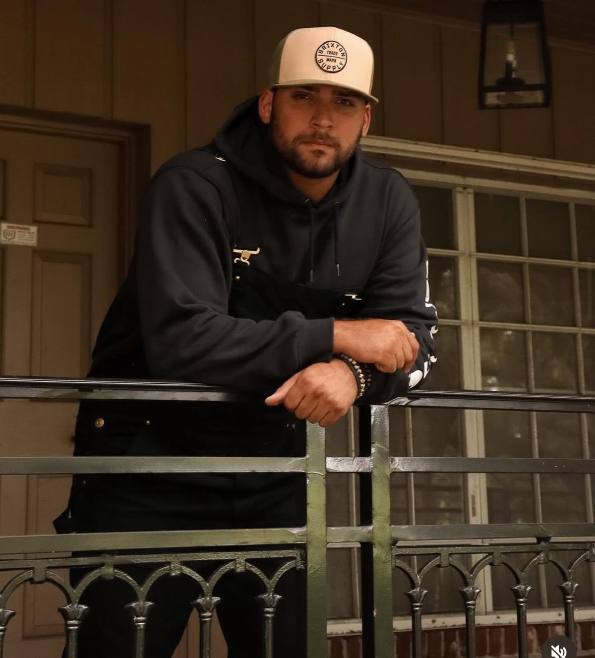 Man in black overalls and black hoodie wearing a beige baseball cap.
