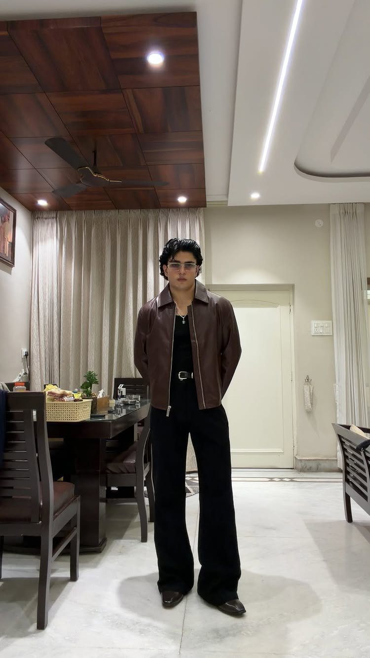 Man wearing a brown leather jacket and wide-leg black trousers in a room.