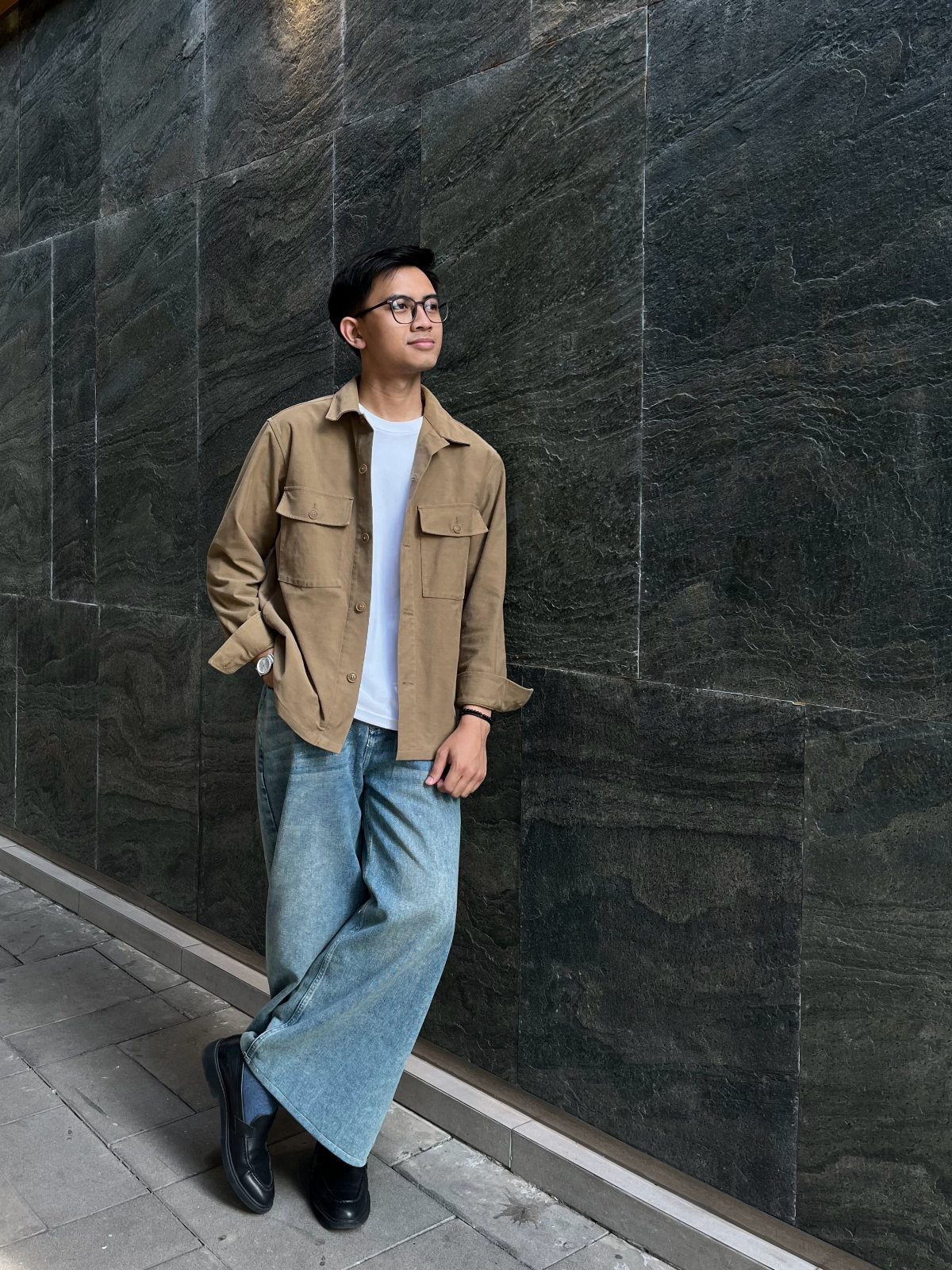 Man wearing a brown jacket, white t-shirt, and wide-leg blue jeans by a wall.