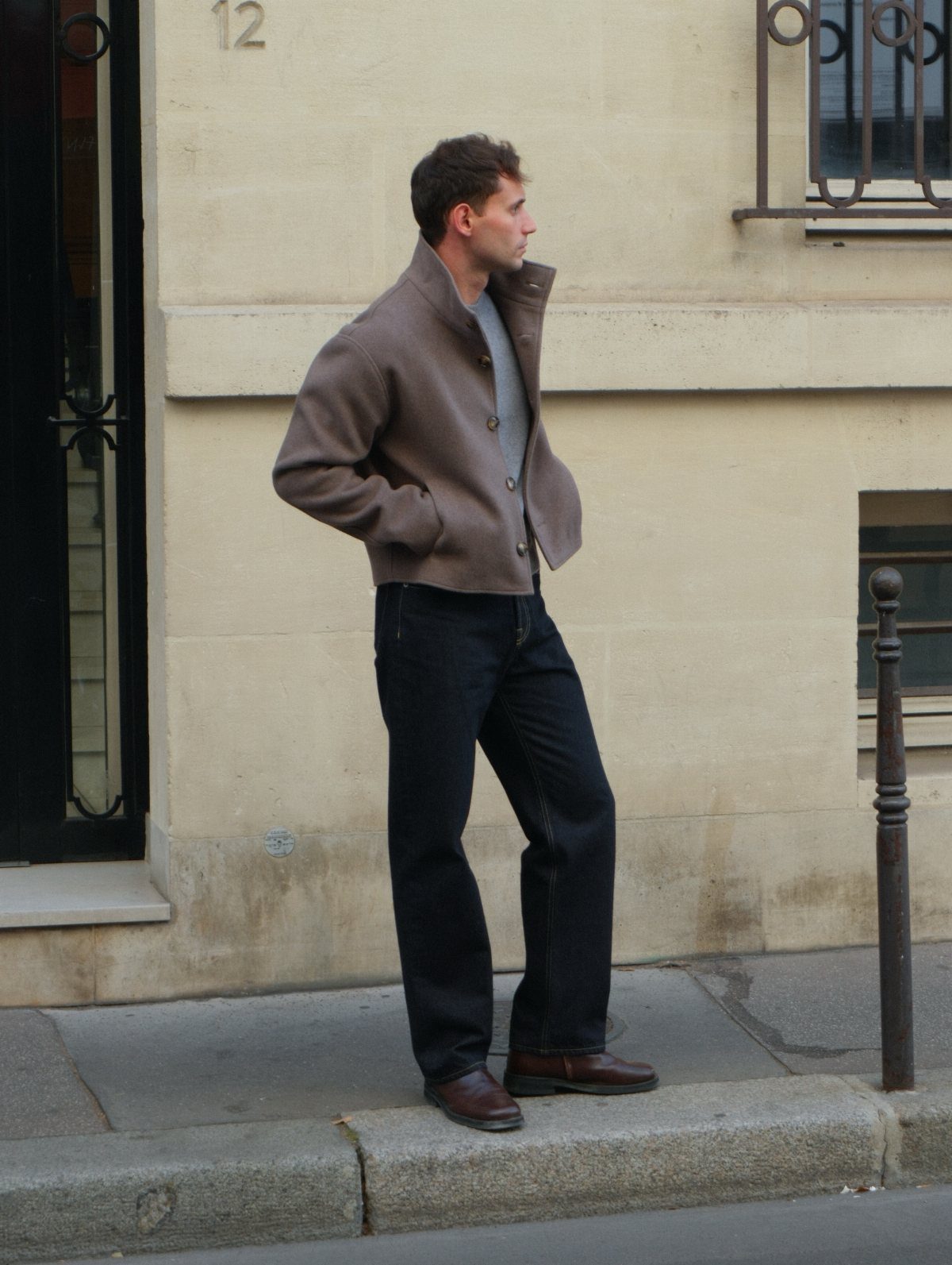 Man wearing a brown jacket and navy jeans standing on a city sidewalk.
