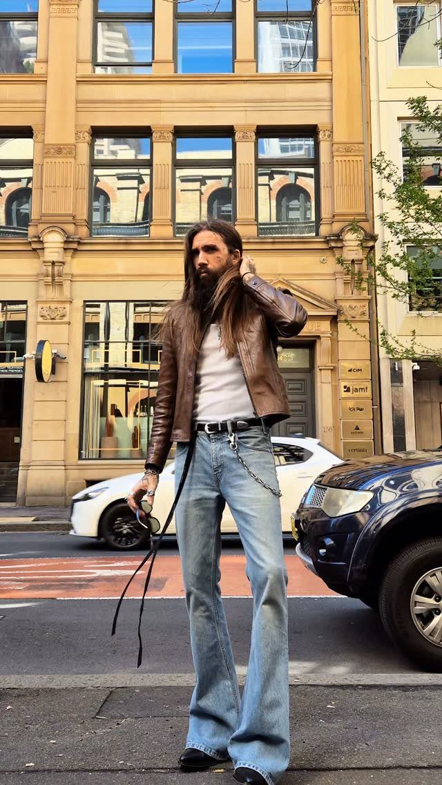 Man wearing a brown leather jacket and light blue flared jeans on city street