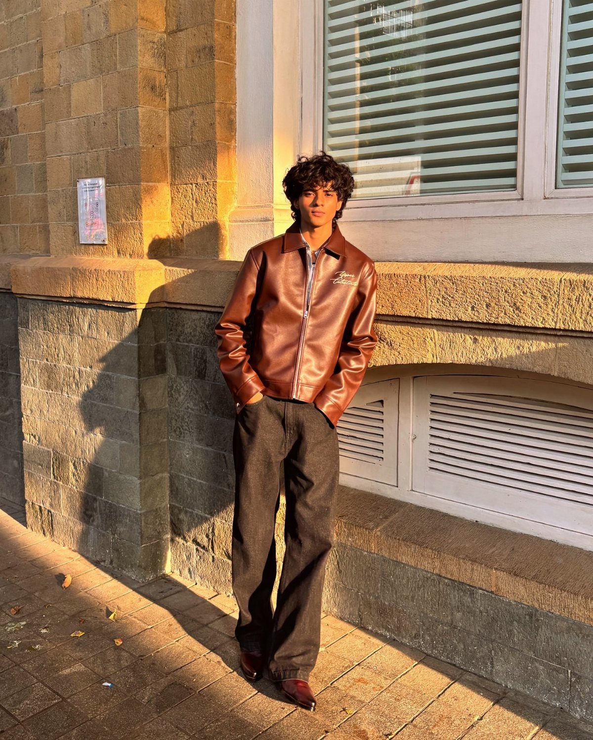 Man wearing a brown leather jacket and dark jeans standing by a stone wall