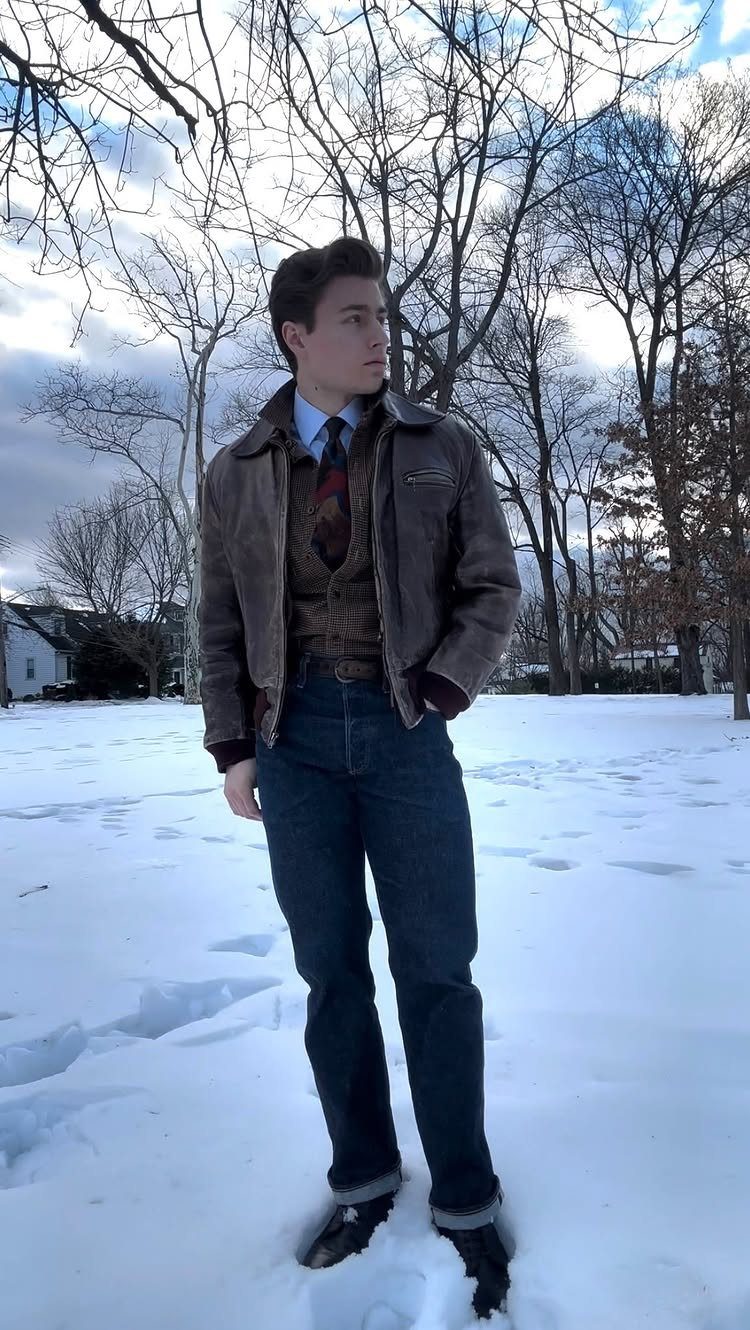Man in brown leather jacket, patterned sweater, and blue jeans standing in snow.