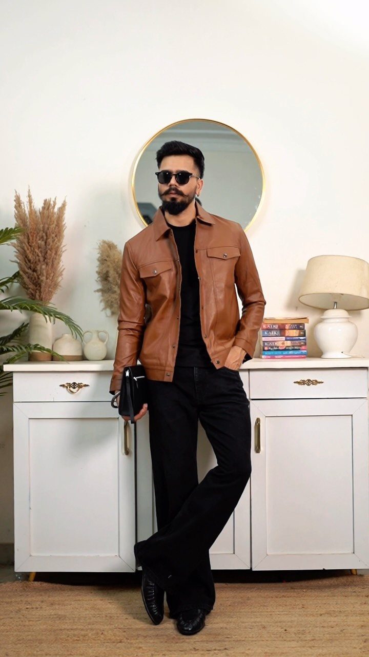 Man in brown leather jacket and black jeans posing in a bright room