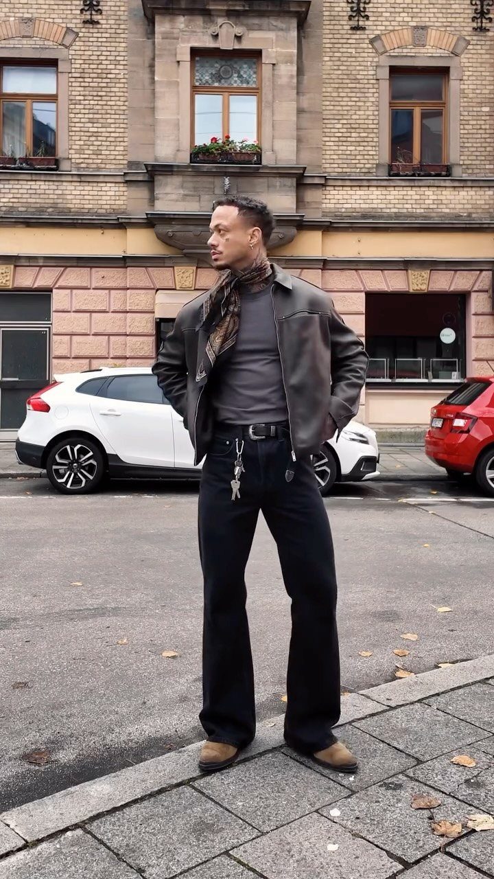 Man wearing brown leather jacket, black wide-leg jeans, and a patterned scarf outdoors.