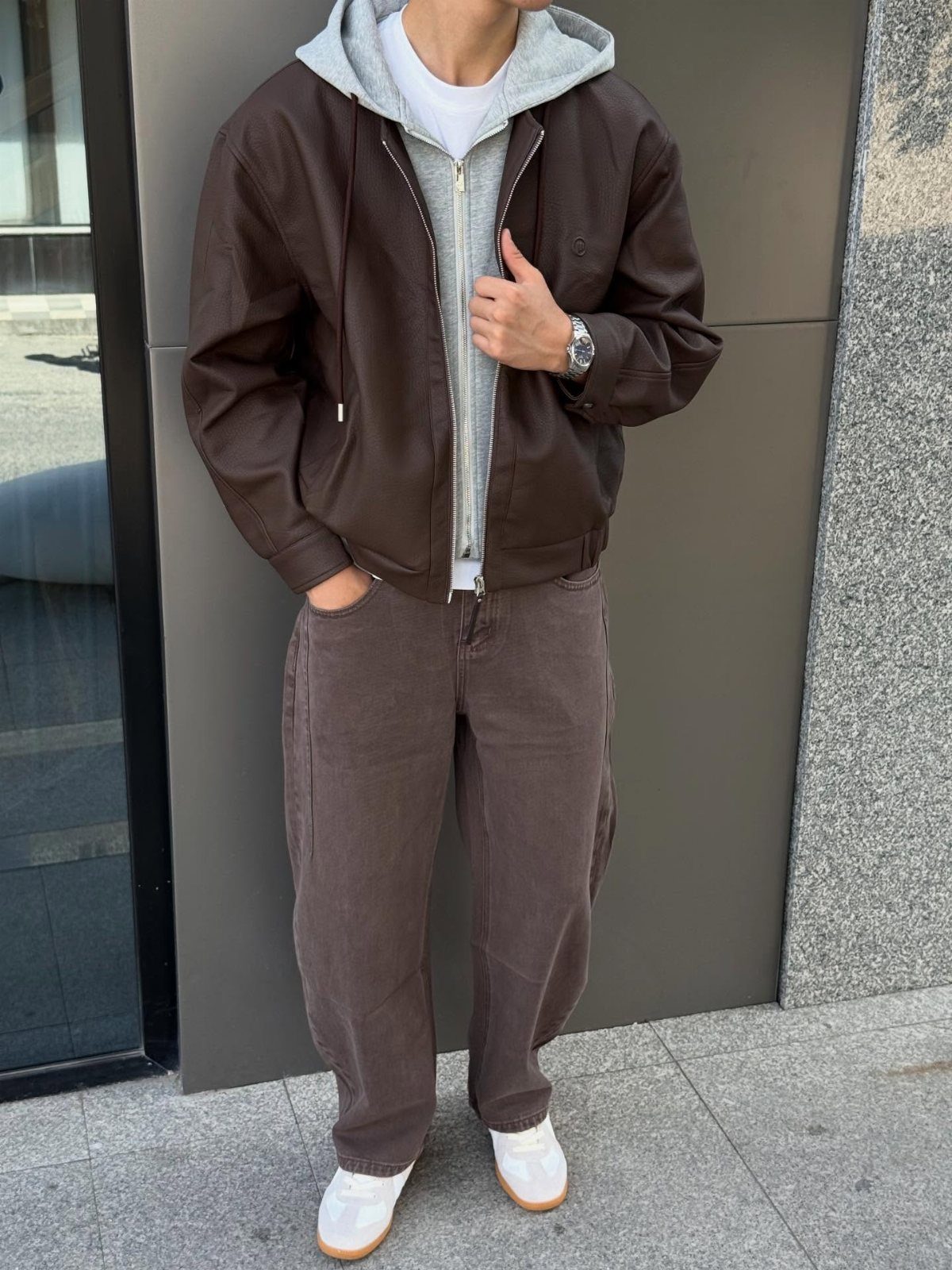 Person wearing a brown leather jacket layered over a grey hoodie and brown trousers.