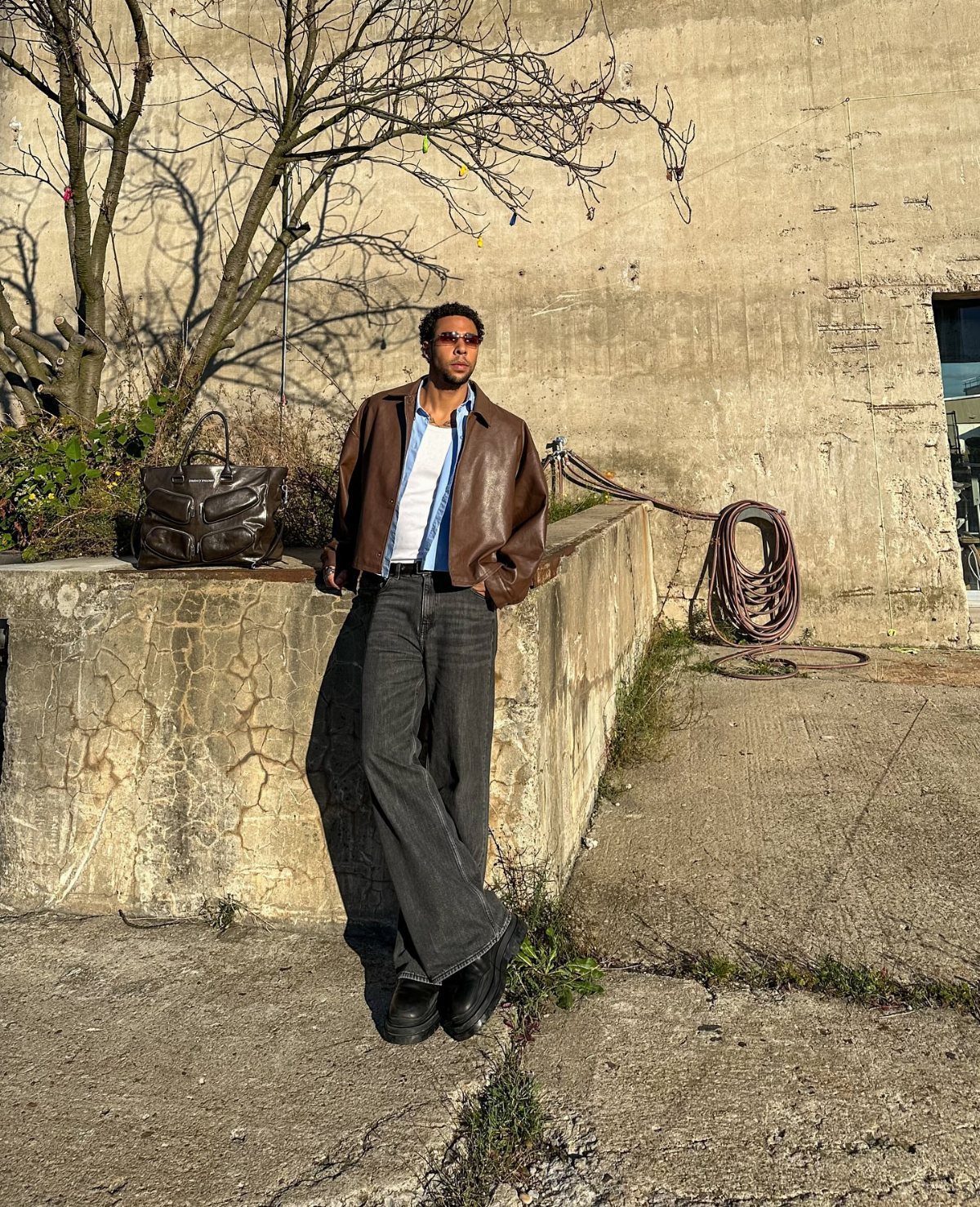 Man wearing a brown leather jacket and wide-leg black trousers standing by a wall