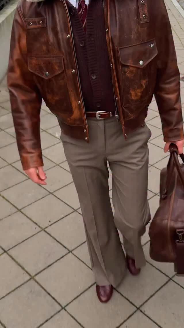 Person wearing a brown leather jacket, brown cardigan, and taupe trousers on pavement.