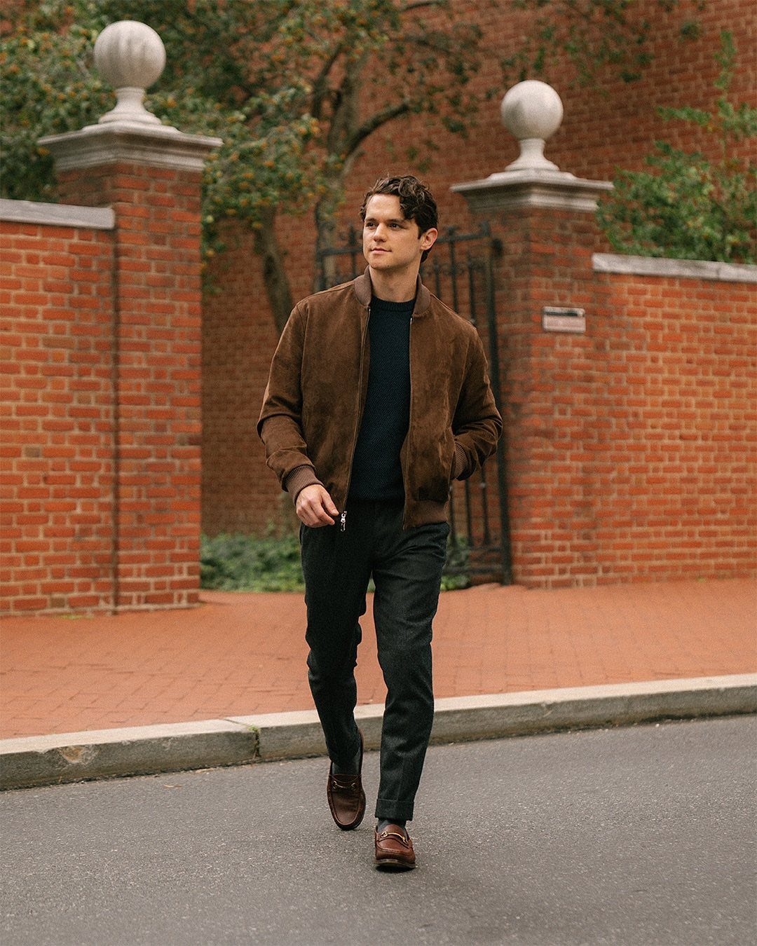 Man wearing a brown suede bomber jacket and dark trousers walking on street