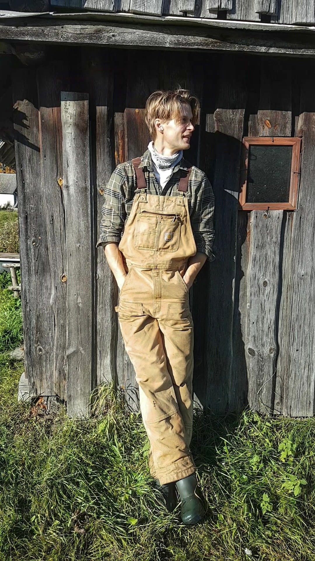 Person wearing brown canvas overalls and a plaid shirt standing by wooden wall