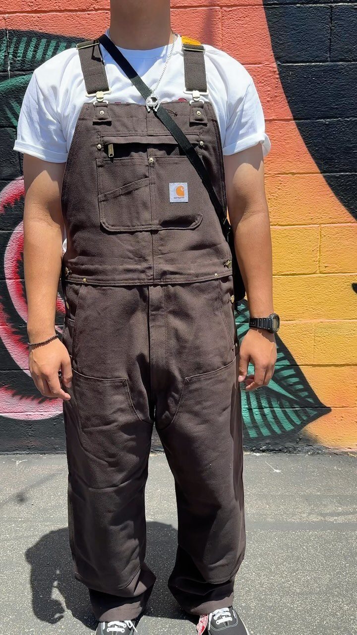 Person wearing brown canvas overalls and a white t-shirt against a graffiti wall