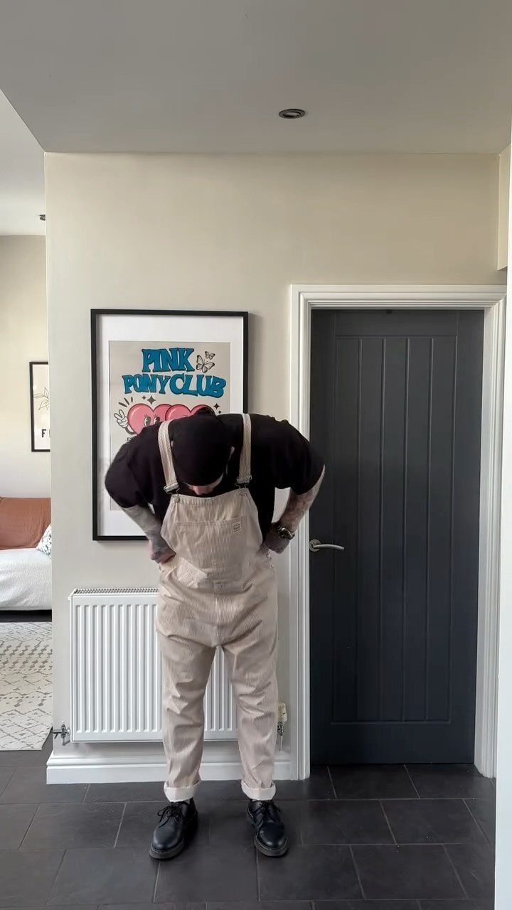 Person wearing light tan overalls and a black t-shirt standing indoors.