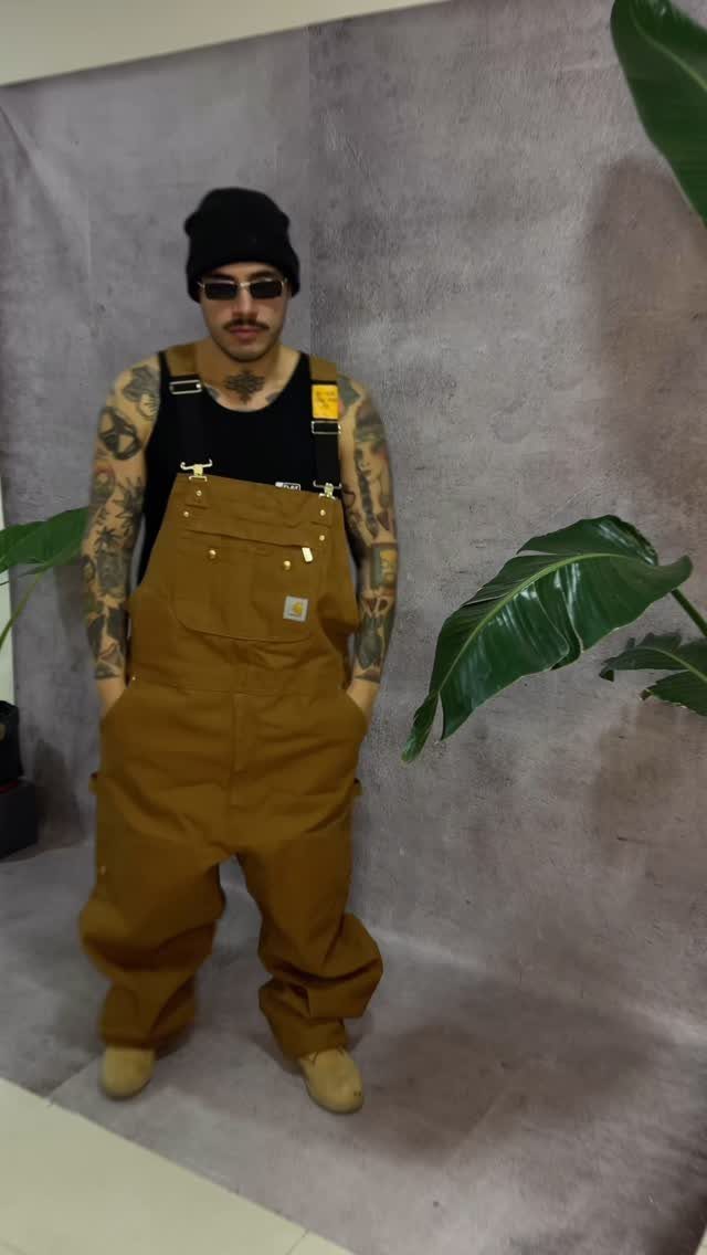 Man wearing brown canvas overalls, black tank top, black beanie, and sunglasses.