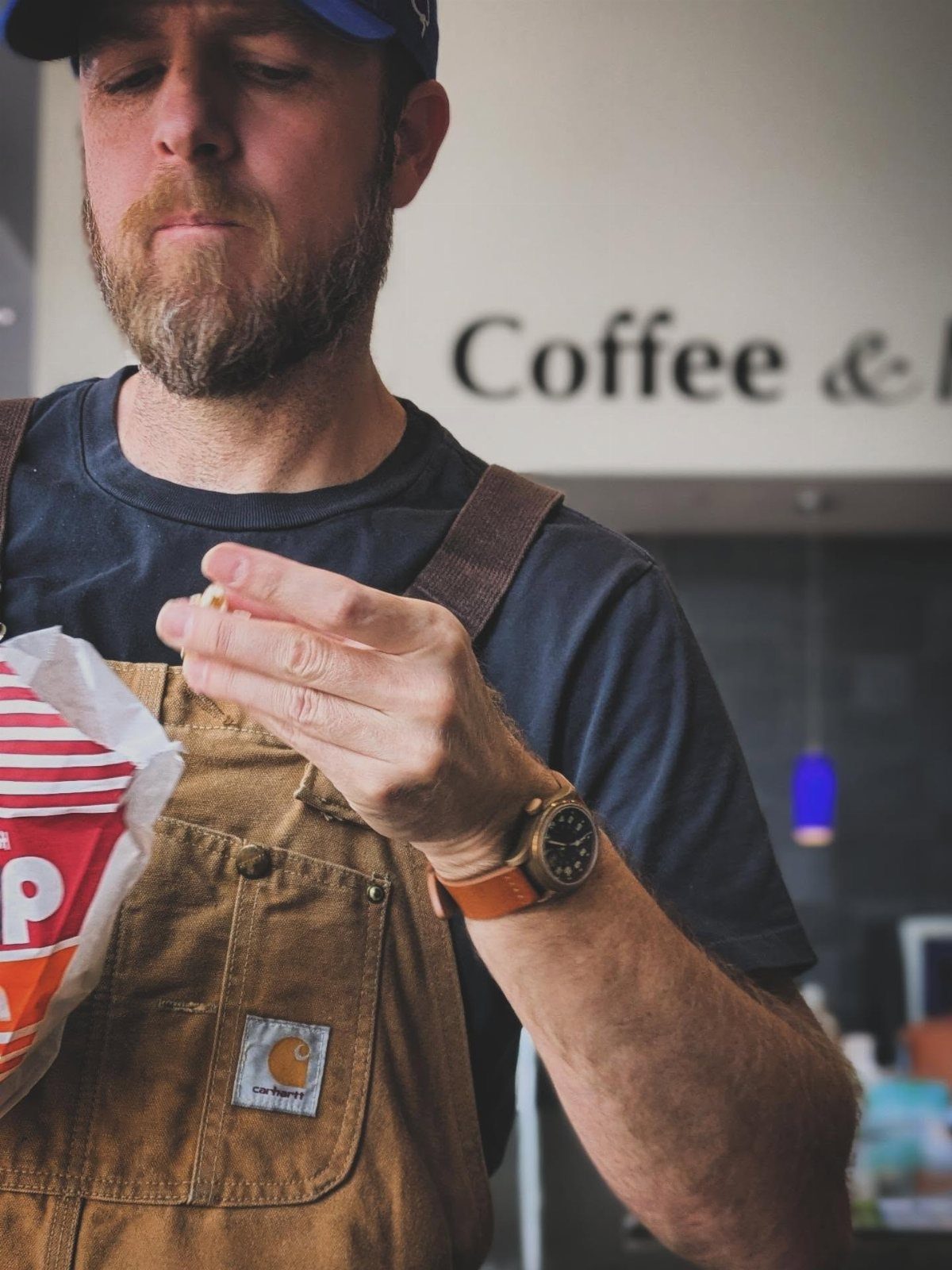 Man in brown canvas overalls and navy t-shirt eating a snack indoors.