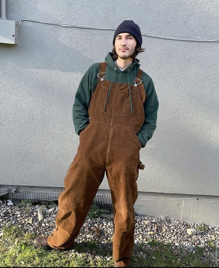 Man wearing brown corduroy overalls, a green hoodie, and a navy blue beanie.