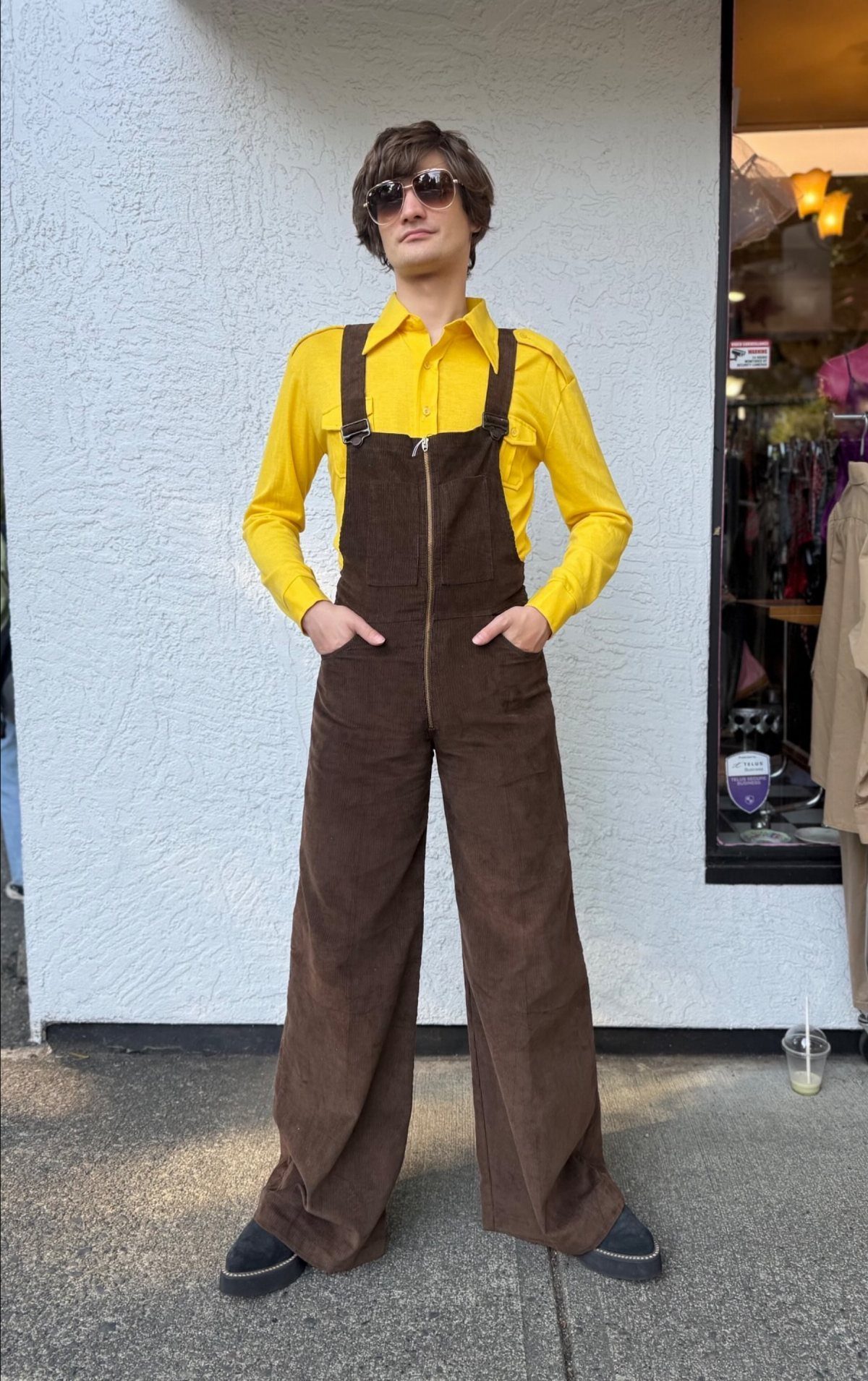 Man wearing brown wide-leg corduroy overalls over a bright yellow button-down shirt.