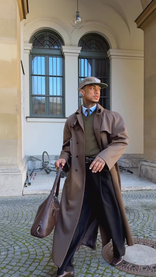 Man in brown trench coat, olive sweater, and black dress pants on cobblestone street