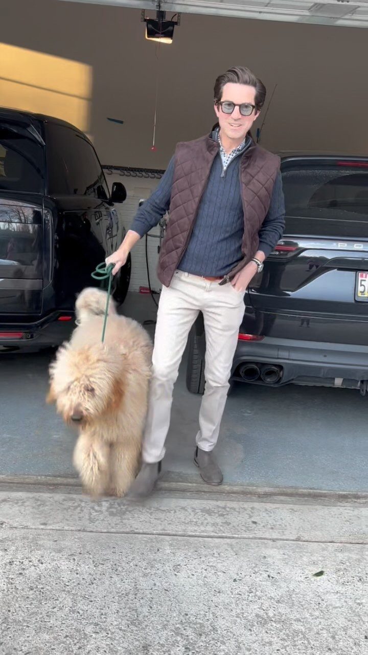 Man in brown quilted vest and beige trousers standing with a fluffy dog.