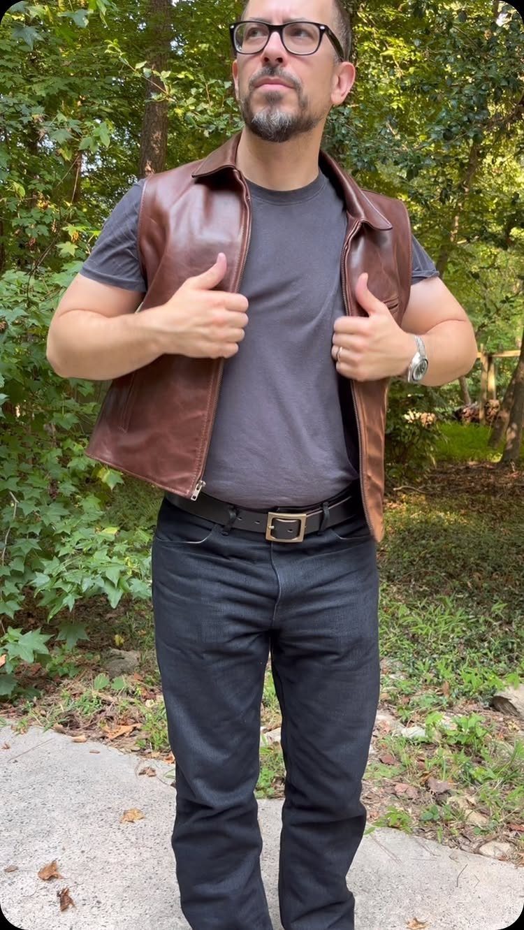 Man wearing a brown leather vest, grey t-shirt, and black jeans outdoors.